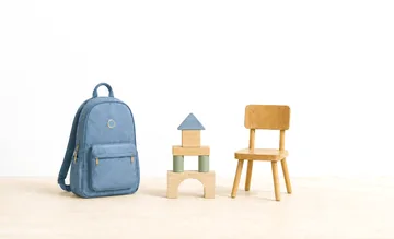 Three things standing on a pale wooden floor against a sunlit wall: a blue backpack, a small tower of wooden blocks, and a child-sized wooden chair